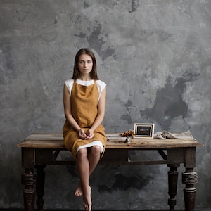 Op de afbeelding: Een vrouw met een bruine linnen schort zit op een houten tafel met een klein ingelijst plaatje en een decoratief object op de tafel. De achtergrond is een getextureerde grijze muur.