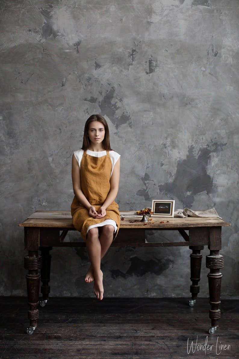 Op de afbeelding: Een vrouw met een bruine linnen schort zit op een houten tafel met een klein ingelijst plaatje en een decoratief object op de tafel. De achtergrond is een getextureerde grijze muur.