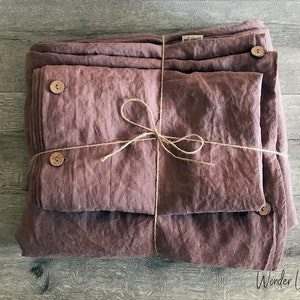  May include: A set of mauve-colored linen bedding, neatly folded and tied with twine. The set includes a duvet cover and pillowcases, secured with wooden buttons. The Wonder Linen logo is visible in the bottom right corner.