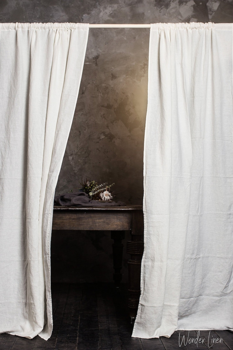Puede incluir: Cortinas de lino blanco cuelgan de una barra delante de una pared gris texturizada. Una mesa de madera con un mantel gris y flores secas se encuentra en el centro de la habitaci&oacute;n.