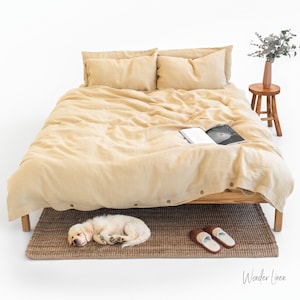 May include: A bed with a light tan linen duvet cover and matching pillows. A small wooden stool holds a vase with greenery. A sleeping puppy rests on a brown woven rug near a pair of brown and white slippers. The text "Wonder Linen" is in the bottom right corner.