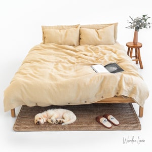  May include: A bed with a light tan linen duvet cover and matching pillows. A small wooden stool holds a vase with greenery. A sleeping puppy rests on a brown woven rug near a pair of brown and white slippers. The text "Wonder Linen" is in the bottom right corner.