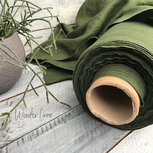 Può includere: Un rotolo di tessuto di lino verde scuro, srotolato su una superficie di legno. Il tessuto è arrotolato attorno a un tubo di cartone. Il testo "Wonder Linen" è visibile sulla superficie.