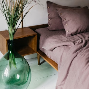 May include: A bedroom scene featuring a bed with mauve-colored linen bedding and pillows with wooden button details. A wooden nightstand and a large green glass vase with tall grass are also visible. The text "Wonder Linen" is at the bottom.