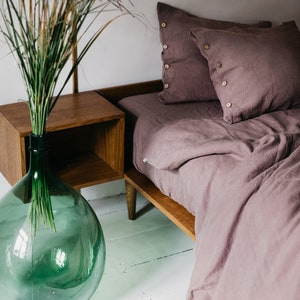  May include: A bedroom scene featuring a bed with mauve-colored linen bedding and pillows with wooden button details. A wooden nightstand and a large green glass vase with tall grass are also visible. The text "Wonder Linen" is at the bottom.