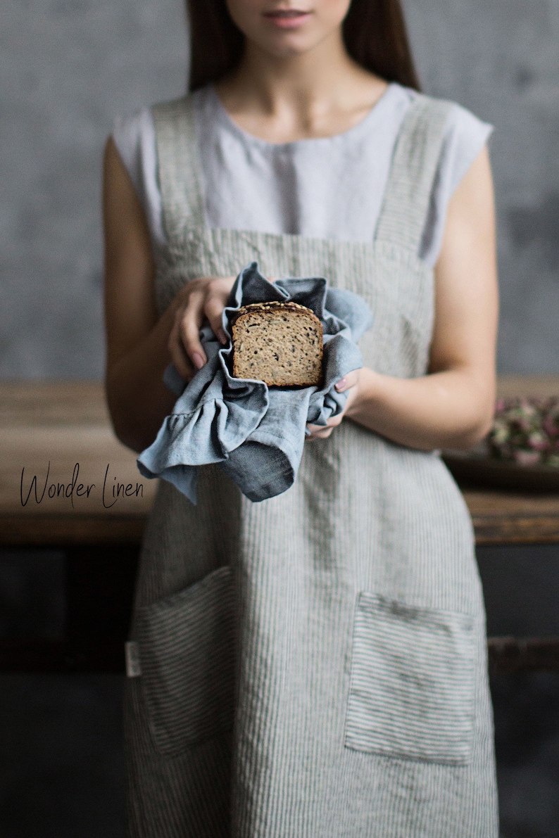 May include: A woman wearing a light blue and white striped linen apron holds a slice of bread wrapped in a blue linen tea towel. The text "Wonder Linen" is visible in the lower left corner of the image.