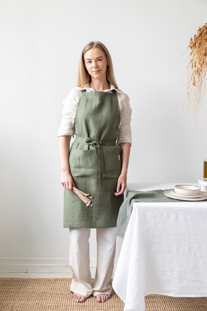 Op de afbeelding: Een vrouw draagt een saliegroene linnen schort met een grote zak. Ze staat voor een witte tafel met een wit tafelkleed en een groene linnen loper.