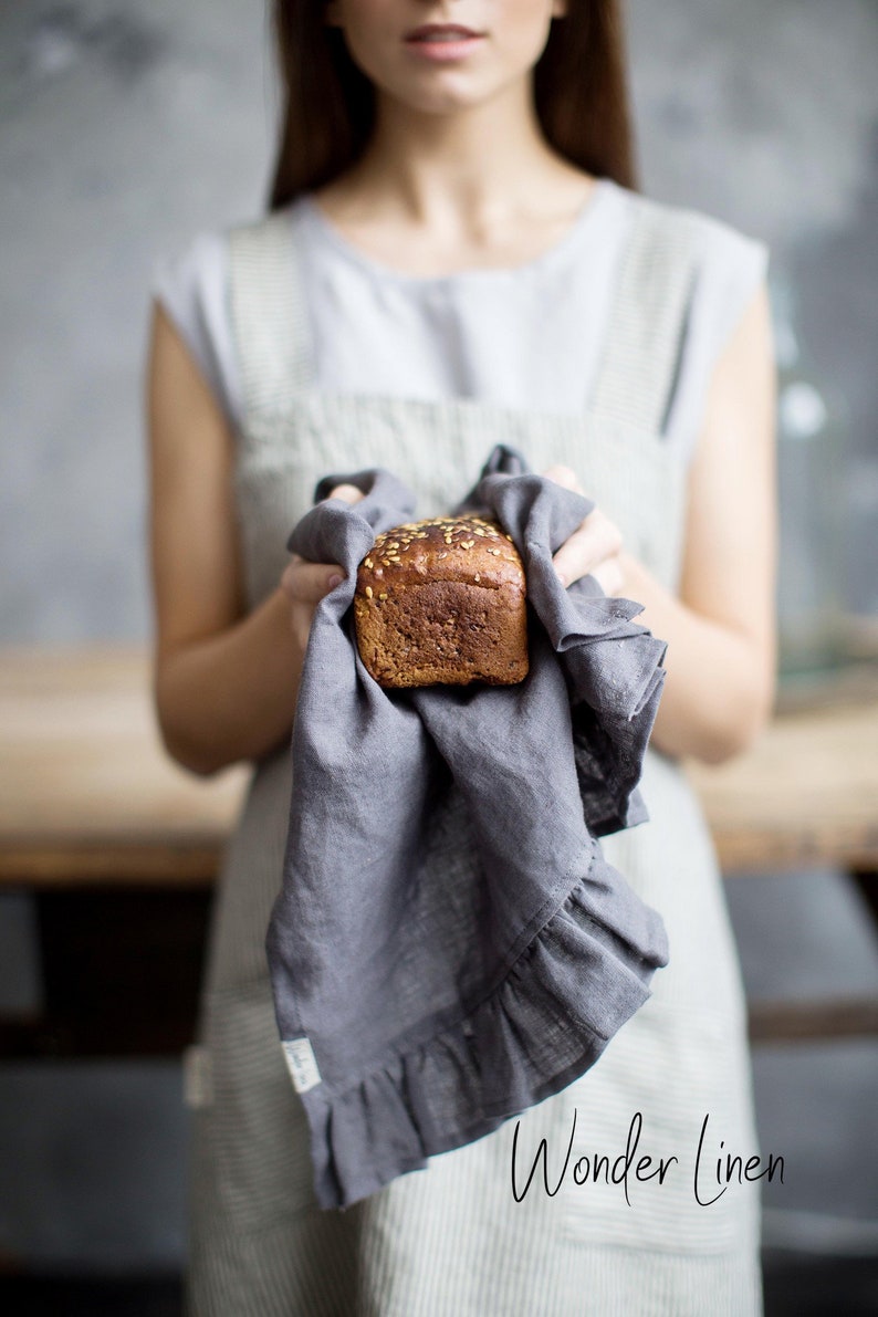 May include: A woman holds a loaf of bread wrapped in a gray linen tea towel with a ruffled edge. The text "Wonder Linen" is visible at the bottom of the image.