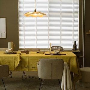 May include: A yellow linen tablecloth with the text "Wonder Linen" on it, set on a dining table with two chairs. The table is set with a pitcher, plates, and a candle. The room has a brown wall and a window with white blinds.