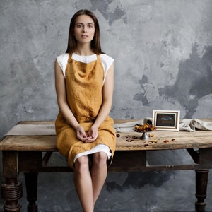 Op de afbeelding: Een vrouw draagt een wit topje en een mosterdgele linnen schort en zit op een houten tafel. De tafel is versierd met gedroogde bloemblaadjes. De achtergrond is een getextureerde grijze muur.