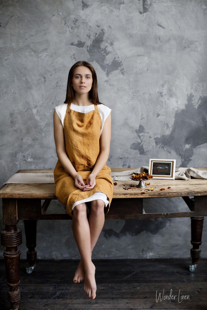 Op de afbeelding: Een vrouw draagt een wit topje en een mosterdgele linnen schort en zit op een houten tafel. De tafel is versierd met gedroogde bloemblaadjes. De achtergrond is een getextureerde grijze muur.