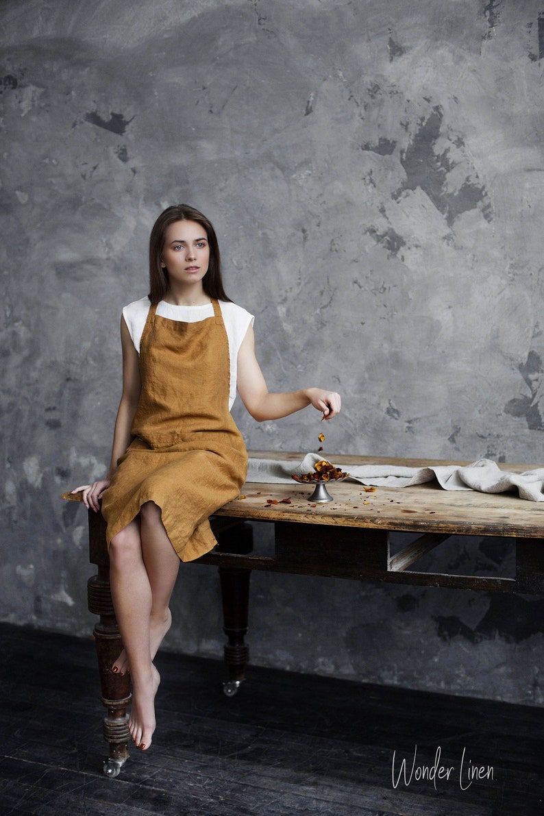 Op de afbeelding: Een vrouw met een bruine linnen schort zit aan een houten tafel met een klein zilveren schaaltje erop. De tafel heeft een rustieke uitstraling en staat op wielen. De achtergrond is een grijze betonnen muur. De tekst "Wonder Linen" is zichtbaar in de rechterbenedenhoek van de afbeelding.