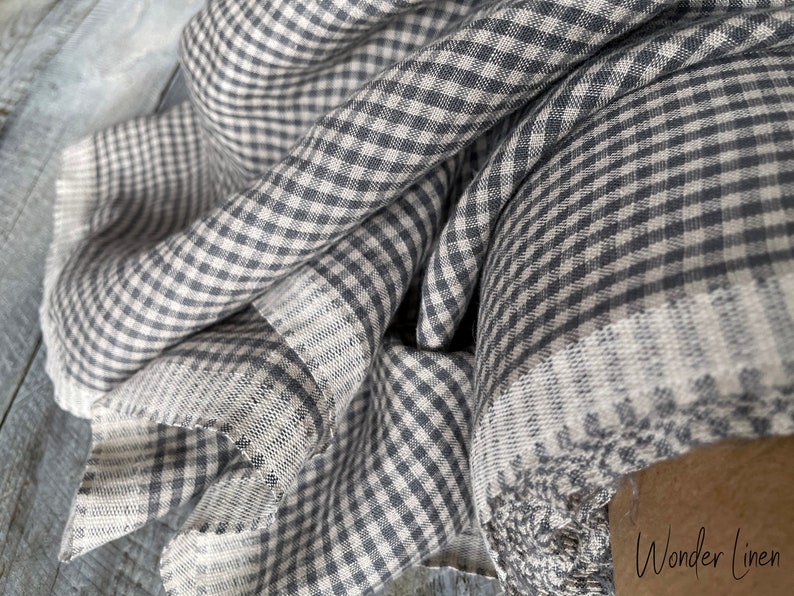 May include: A close-up of a gray and white gingham fabric. The fabric is folded and has a soft, textured feel. The fabric is made of linen and is perfect for sewing projects.
