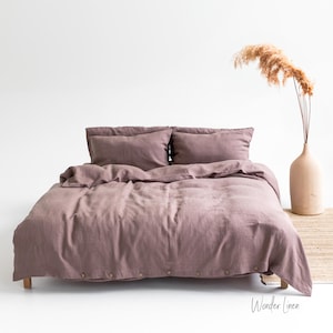 May include: A bed with a mauve linen duvet cover and two matching pillows. The bed is on a wooden frame. A beige vase with dried flowers sits on a small wooden table. The text "Wonder Linen" is visible on the bottom right.