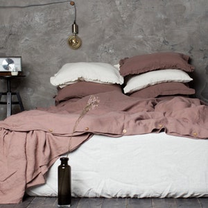  May include: A bed with white and mauve linen bedding. The bed is made with a white fitted sheet, a mauve duvet cover with wooden buttons, and several pillows in white and mauve. A dark vase with dried flowers is in the foreground.