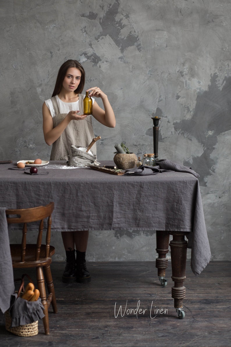 May include: A woman wearing a linen apron stands in front of a table with a grey linen tablecloth. She is holding a brown glass bottle. There are various kitchen items on the table, including a mortar and pestle, a wooden spoon, and a bowl of flour. The text "Wonder Linen" is visible in the bottom right corner of the image.