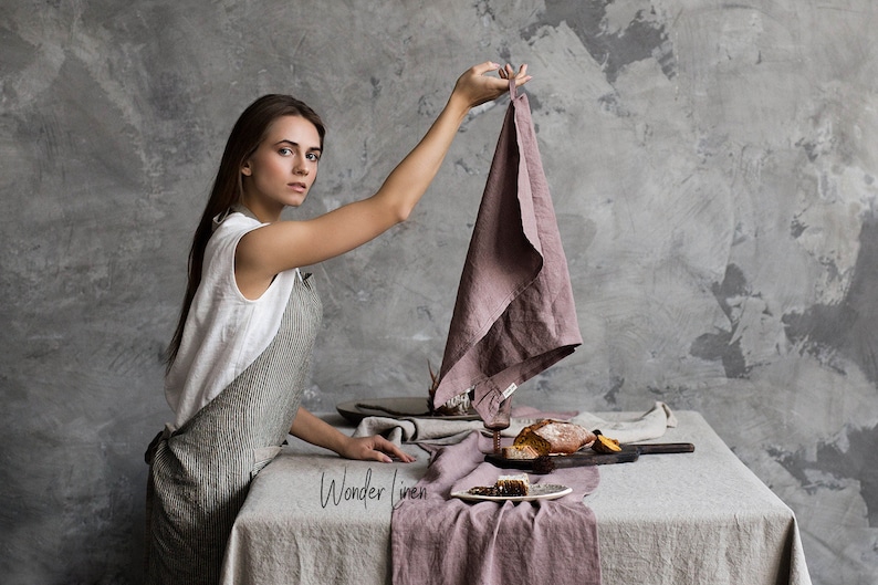 May include: A woman in a white sleeveless shirt and a gray linen apron stands at a table set with a linen tablecloth and a pink linen runner. The table is decorated with a loaf of bread, fruit, and a glass of wine. The woman is holding a pink linen tea towel. The text "Wonder Linen" is visible on the table.