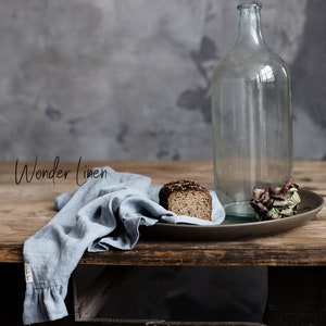May include: A rustic wooden table with a blue linen cloth, a clear glass bottle, a slice of dark bread on a brown plate, and a dried flower.