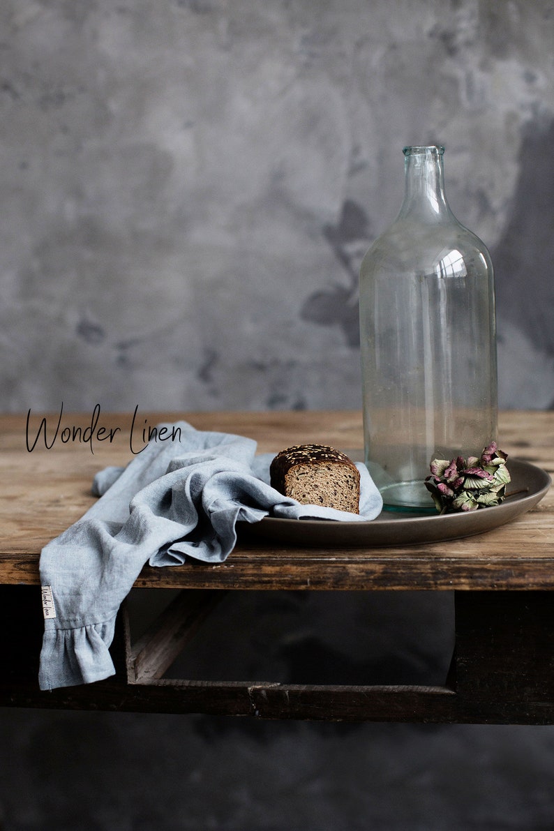 May include: A rustic wooden table with a blue linen cloth, a clear glass bottle, a slice of dark bread on a brown plate, and a dried flower.
