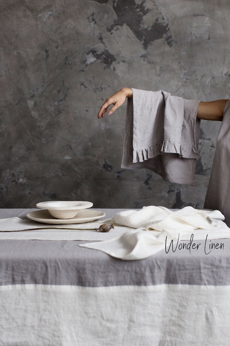 May include: A gray linen tablecloth with a white border. The tablecloth is set with a white ceramic bowl and a white linen napkin. The text "Wonder Linen" is visible in the lower right corner of the image.