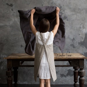 May include: A woman wearing a white linen dress and a beige linen apron stands in front of a grey concrete wall. She is holding a grey linen sheet in her hands. The text "Wonder Linen" is visible in the lower right corner of the image.