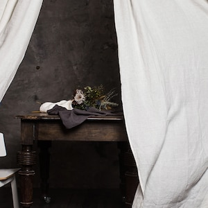 May include: A dark wooden table with a brown cloth and a bouquet of dried flowers on top. The table is on wheels and is positioned in front of a grey wall. Two white linen curtains are on either side of the table.