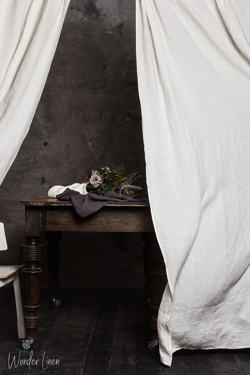 May include: A dark wooden table with a brown cloth and a bouquet of dried flowers on top. The table is on wheels and is positioned in front of a grey wall. Two white linen curtains are on either side of the table.