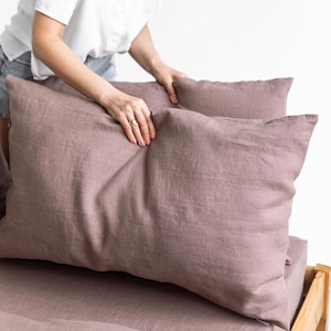 May include: Two mauve linen pillowcases are being placed on a bed. The pillowcases are made of a soft, natural fabric and have a subtle sheen.