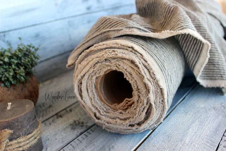 K&ouml;nnte beinhalten: Eine Rolle aus nat&uuml;rlichem Leinenstoff mit schwarzen Streifen. Der Stoff ist aufgerollt und hat eine rohe Kante. Der Stoff liegt auf einer Holzoberfl&auml;che.