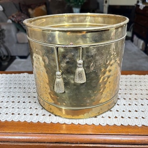 Peut inclure: Un seau cylindrique en laiton présentant une texture martelée sur le corps principal et un bord lisse. L'extérieur est orné d'une bordure horizontale en forme de corde et de deux pompons en laiton suspendus à un détail de corde assorti.