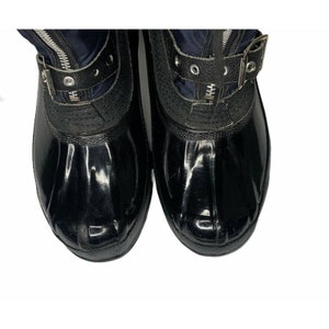 K&ouml;nnte beinhalten: Ein Paar schwarze Gummistiefel mit einem dunkelblauen Nylon-Obermaterial. Die Stiefel haben einen Rei&szlig;verschluss an der Vorderseite und einen Schnallenriemen &uuml;ber dem Obermaterial.