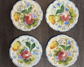 Könnte beinhalten: Vier dekorative Keramikteller mit gewellten Rändern. Jeder Teller zeigt ein zentrales Rosendesign in Rot und Rosa, umgeben von grünen Blättern, blauen Blumen und einer gelben Frucht. Ein blauer Rand säumt die Teller.