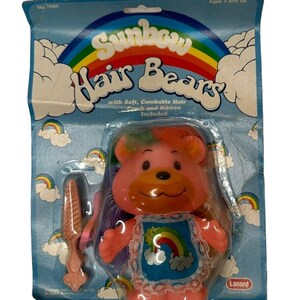 Puede incluir: Un oso de juguete de plástico rosa con pelo de color arcoíris, que lleva un vestido azul con un diseño de arcoíris y nubes. El oso viene con un peine y una cinta. El embalaje dice "Sunbow Hair Bears" y "No. 7080".