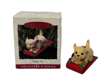 Könnte beinhalten: Ein weißer Welpen-Anhänger auf einem roten Schlitten mit einer goldenen Glocke. Der Anhänger ist Teil der Hallmark Keepsake Ornament Collector's Series und trägt auf der Schachtel den Schriftzug "Puppy Love".