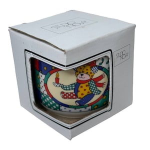 May include: A white ceramic mug with a colorful patchwork design featuring a teddy bear holding a candle. The mug is in a white box with the text "Studio Nova" printed on the side.