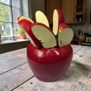 Może przedstawiać: Dekoracyjny czerwony uchwyt na noże w kształcie jabłka z sześcioma nożami w kształcie plasterków jabłka. Noże mają czerwone ostrza i białawe krawędzie. Uchwyt stoi na drewnianym stole.