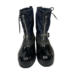 K&ouml;nnte beinhalten: Ein Paar schwarze und marineblaue Winterstiefel mit einem Rei&szlig;verschluss vorne und einem Schnallenriemen. Die Stiefel haben eine gl&auml;nzende schwarze Gummisohle.