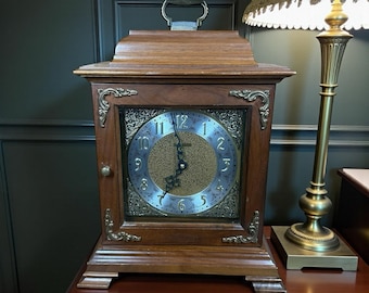 Può includere: Un orologio da mensola in legno di forma rettangolare, con una maniglia sulla parte superiore. Il quadrante ha un centro dorato e numeri argentati. Ornamenti dorati decorano gli angoli. L'orologio è posto su una superficie di legno scuro.
