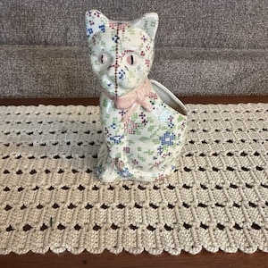 May include: A ceramic cat-shaped planter with a floral cross-stitch pattern in blue, green, and red. The cat has a pink bow and a stitched seam down the middle. The planter sits on a cream-coloured crocheted table runner.