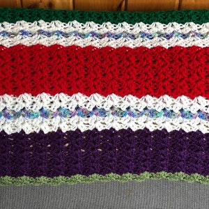 May include: A multi-colored crocheted blanket with stripes of green, red, white, purple, and green. The blanket is made with a textured stitch.