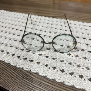 Könnte beinhalten: Runde Brille mit Drahtgestell und klaren Gläsern. Die Fassungen sind dunkelmetallfarben, und die Bügel biegen sich über die Ohren. Die Brille liegt auf einer weißen, gehäkelten Tischdecke, mit einer Holzoberfläche im Hintergrund.