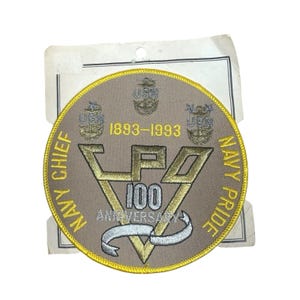 May include: A circular, tan patch with gold and silver embroidery. The patch reads "Navy Chief Navy Pride" and "100 Anniversary" with the years 1893-1993. The patch has a gold border and the letters "CPO" in gold.