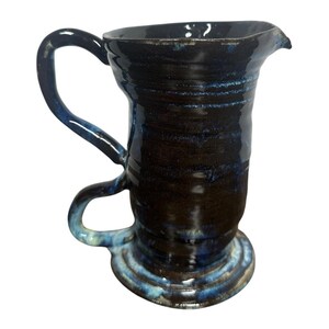 May include: A handmade ceramic pitcher with a dark blue and brown glaze. The pitcher has a curved handle and a spout for pouring. The base and handle have a lighter blue accent. The pitcher is a unique piece of pottery.