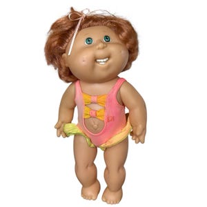 May include: A vintage Cabbage Patch Kid doll with red hair and blue eyes. The doll is wearing a pink and yellow swimsuit with bow accents and a ruffled skirt. A pink bow is in the doll's hair. The doll is standing and smiling.