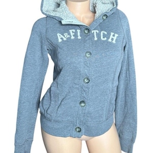 May include: Gray hooded sweatshirt with a button front and a sherpa lined hood. The sweatshirt has the text "A&FITCH" embroidered on the front in white.