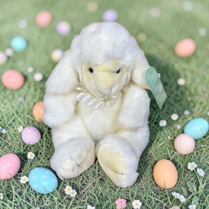 May include: A plush, cream-colored bunny with a beige and white striped bow, sitting on green grass. Pastel Easter eggs and small white flowers are scattered around the bunny, creating a festive scene. A green tag is attached to the bunny.