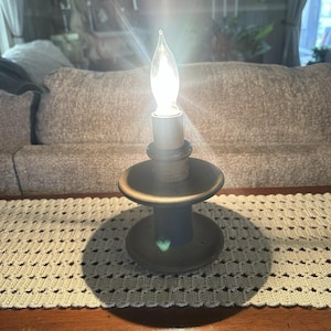 May include: A lit candle in a decorative holder. The candle has a flame-shaped bulb. The holder is dark brown with a round base and a smaller, circular top. The candle sits on a table with a cream-coloured, crocheted runner.