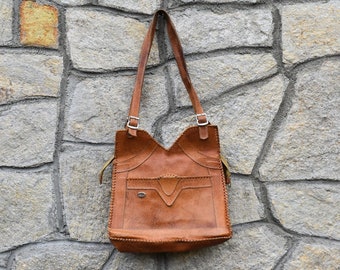 May include: A brown leather tote bag with decorative stitching and a flap closure. The bag is hanging on a stone wall.
