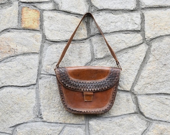 May include: A brown leather shoulder bag with a woven detail on the flap. The bag is hanging on a stone wall.