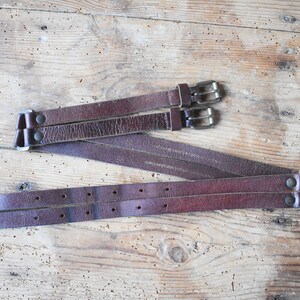 May include: Three brown leather belts with silver buckles. The belts are arranged in a criss-cross pattern on a wooden surface.